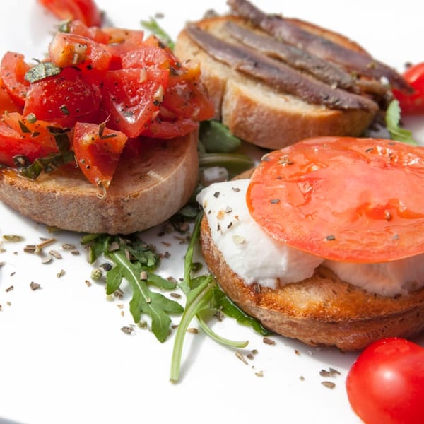 Three bruschetta appetizers on a white plate: tomato, anchovy, and mozzarella with tomato.