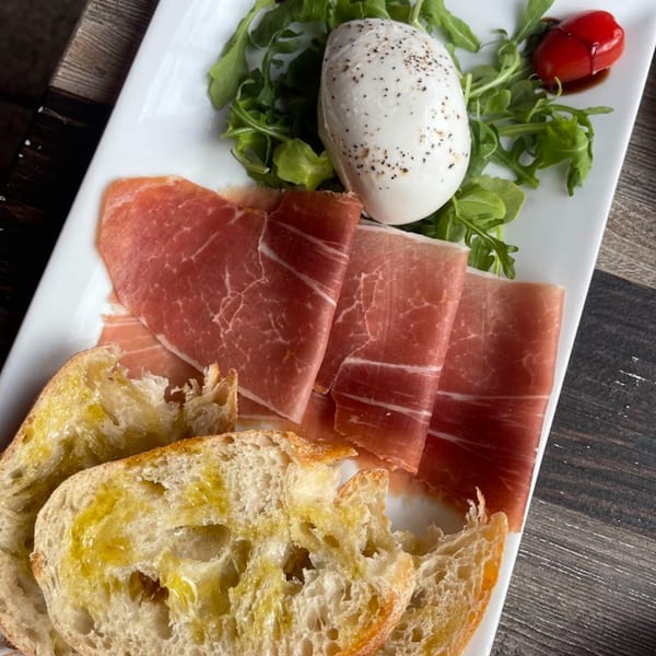 A white plate with prosciutto, burrata cheese, arugula, cherry tomato, and olive oil bread.