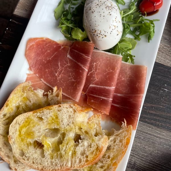 Menomale's prosciutto, burrata, arugula, and cherry tomatoes with olive oil-drizzled bread on a white plate.