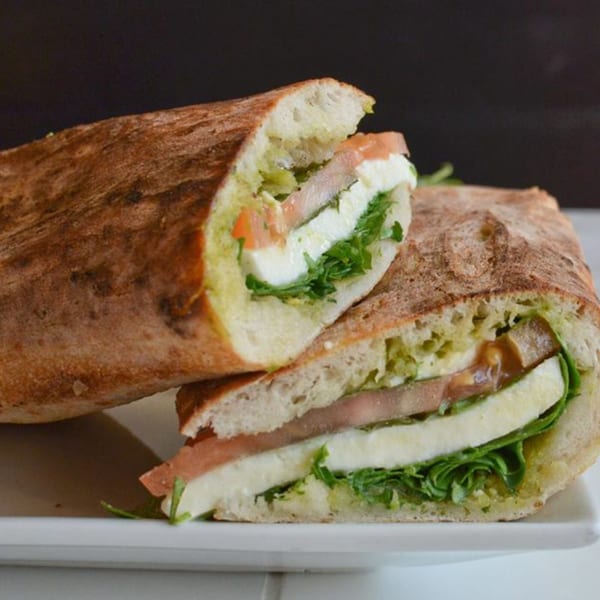 A sliced Italian sandwich with pesto, prosciutto, mozzarella, and arugula on a white plate.