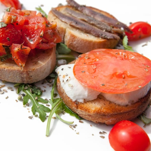 Three bruschetta appetizers on a white plate, topped with tomatoes, anchovies, and mozzarella.