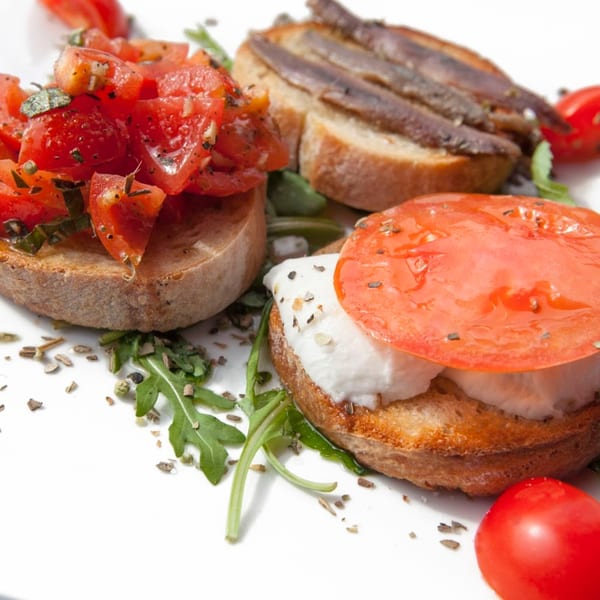 Three bruschetta appetizers: tomato, anchovy, and mozzarella with tomato, on a white plate.
