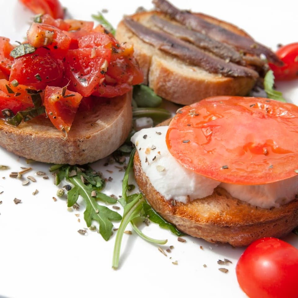 Three bruschetta appetizers: tomato, anchovy, and mozzarella with tomato, on a white plate.