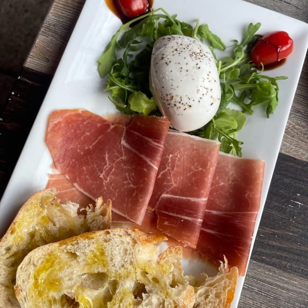 A white rectangular plate holds prosciutto, burrata, arugula, cherry tomatoes, and olive oil-drizzled bread.