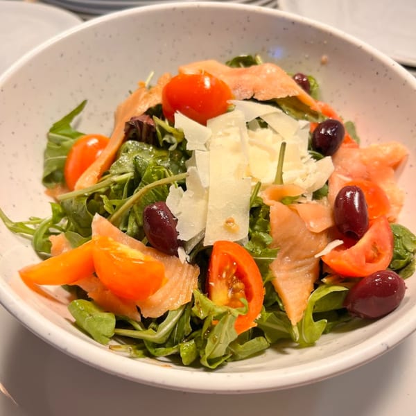 A vibrant salad with arugula, cherry tomatoes, olives, smoked salmon, and shaved parmesan in a white bowl.