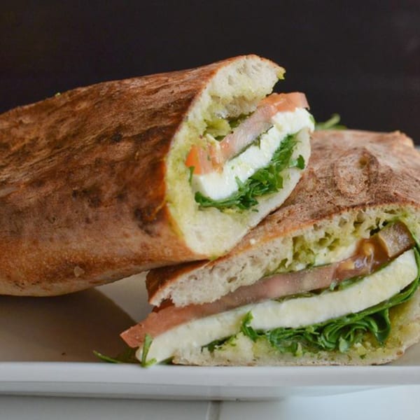 Two halves of a Menomale sandwich with pesto, mozzarella, prosciutto, and arugula on a white plate.